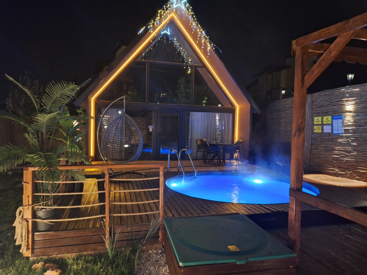 A modern cabin exterior is showcased at night, featuring large glass windows framed with soft lighting. A hot tub is positioned on a wooden deck surrounded by greenery. A hanging chair and dining set invite relaxation in the outdoor space, which is gently illuminated.