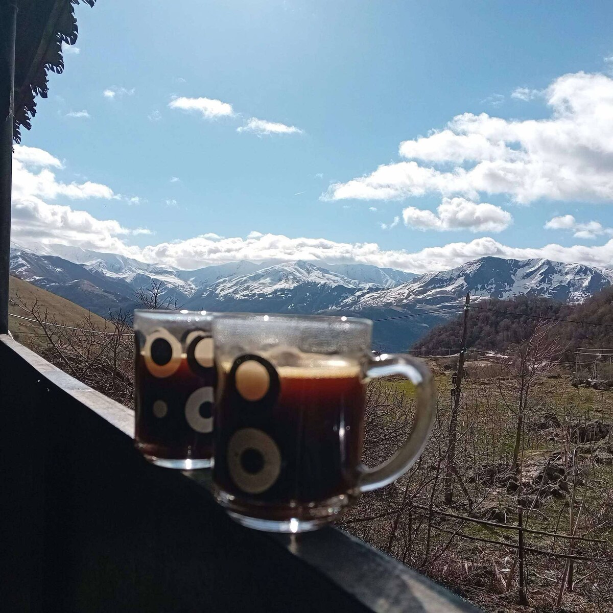 Two clear glass mugs filled with dark coffee are positioned on a railing, overlooking a panoramic view of mountains. Snow-capped peaks stand against a backdrop of blue sky with scattered white clouds.
