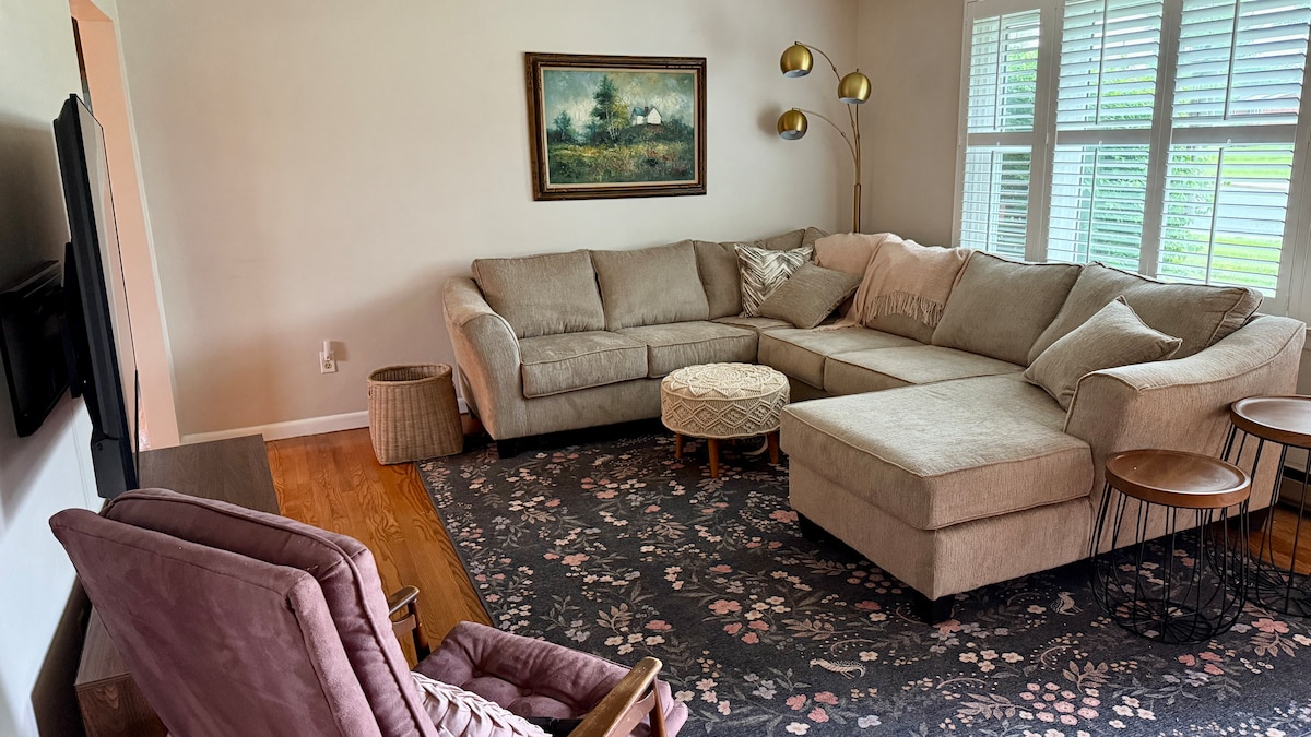 A spacious living area features a comfortable sectional sofa in muted tones, complemented by a round ottoman. Light streams through multiple windows adorned with blinds. A soft area rug with floral patterns provides warmth, while a tall floor lamp adds illumination beside the seating.