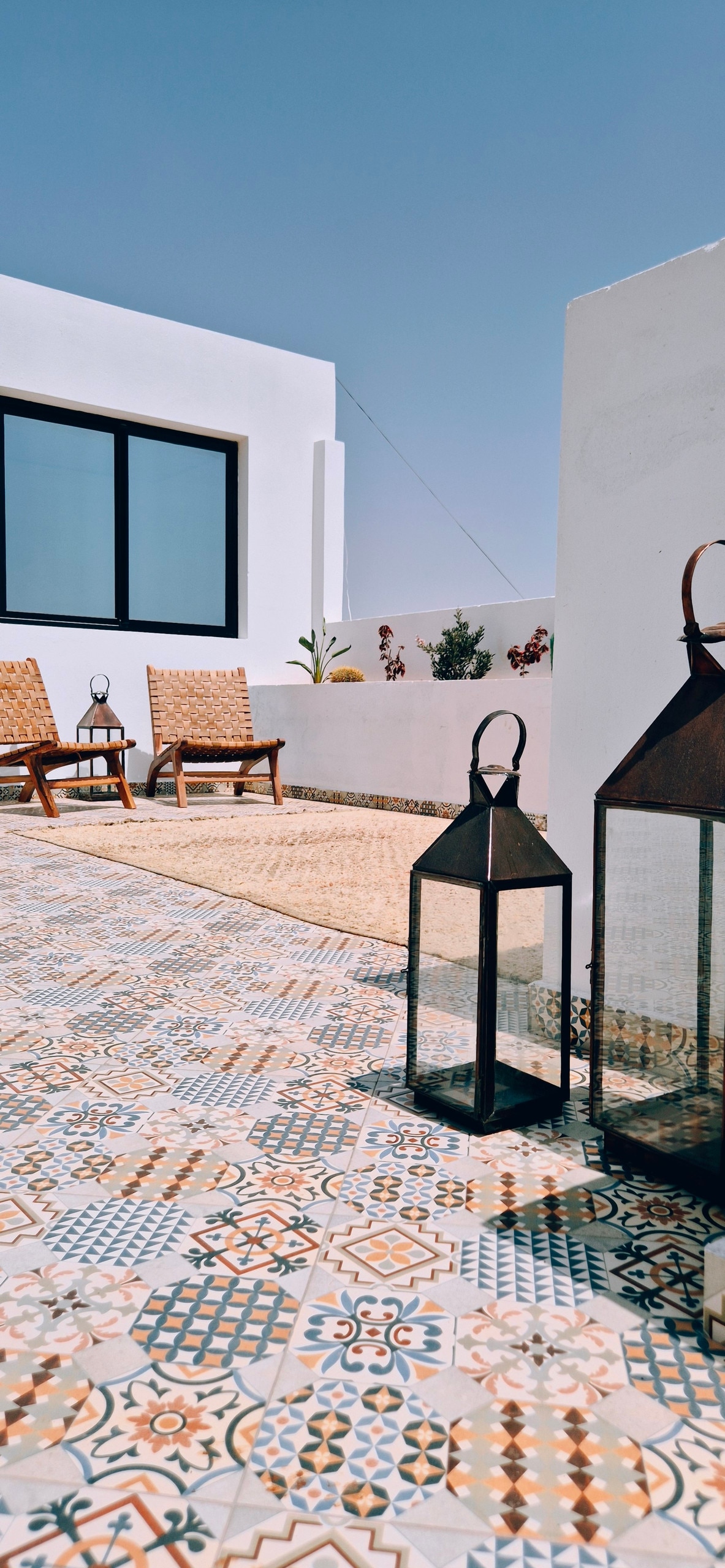 A sunny outdoor terrace is highlighted by two woven chairs positioned on colorful tiled flooring. Decorative lanterns flank the space, adding a touch of character. A large window provides a view of the surrounding area, and greenery is visible in the background.