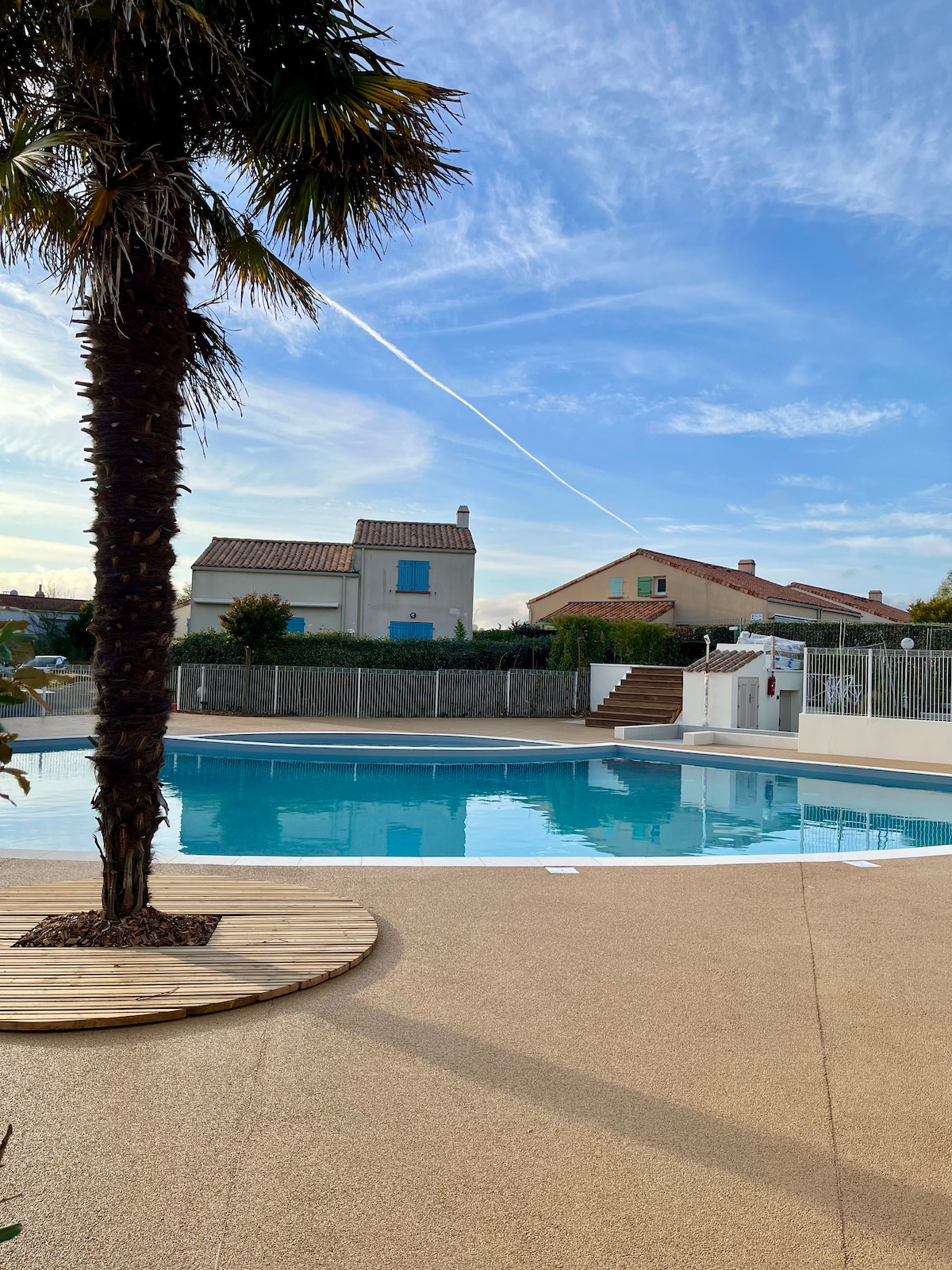 A shared swimming pool is depicted within a residential area, surrounded by a protective fence. A palm tree stands next to a wooden deck adjacent to the pool. The sky is clear, featuring light cloud cover, while buildings are visible in the background.