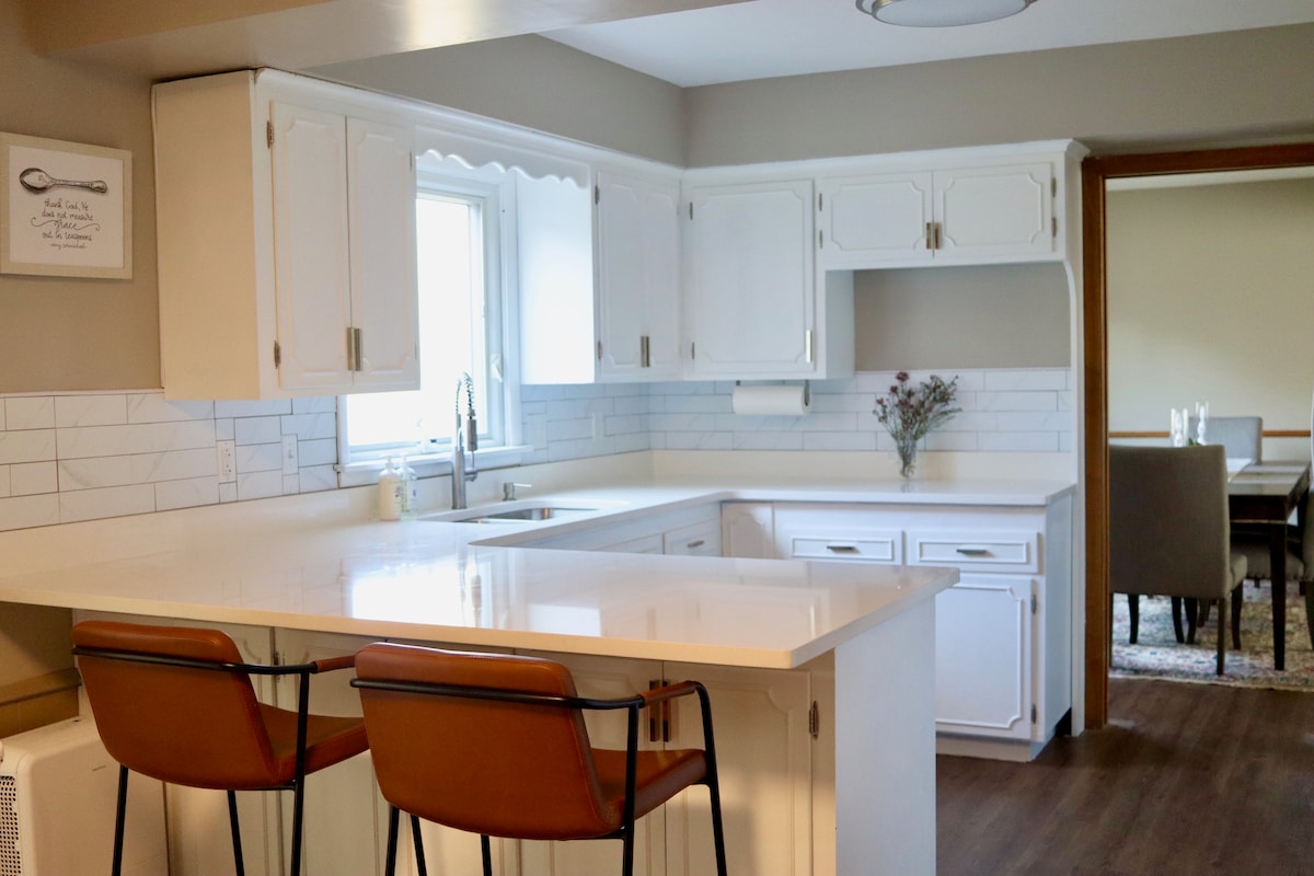 A well-appointed kitchen features white cabinetry and a spacious countertop. Two high-backed stools invite seating at the island. Bright natural light enters through a window above the sink, highlighting the clean, modern lines and subtle tile backsplash.