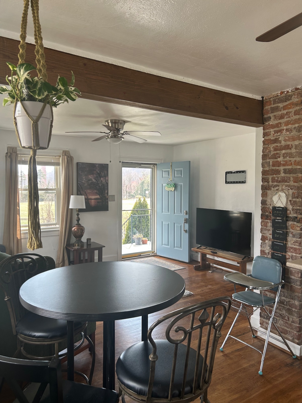 A dining area features a round black table surrounded by four chairs, with a ceiling fan above. A blue front door leads outside, and a small TV is positioned on a stand next to a brick accent wall. Natural light enters through the nearby window.