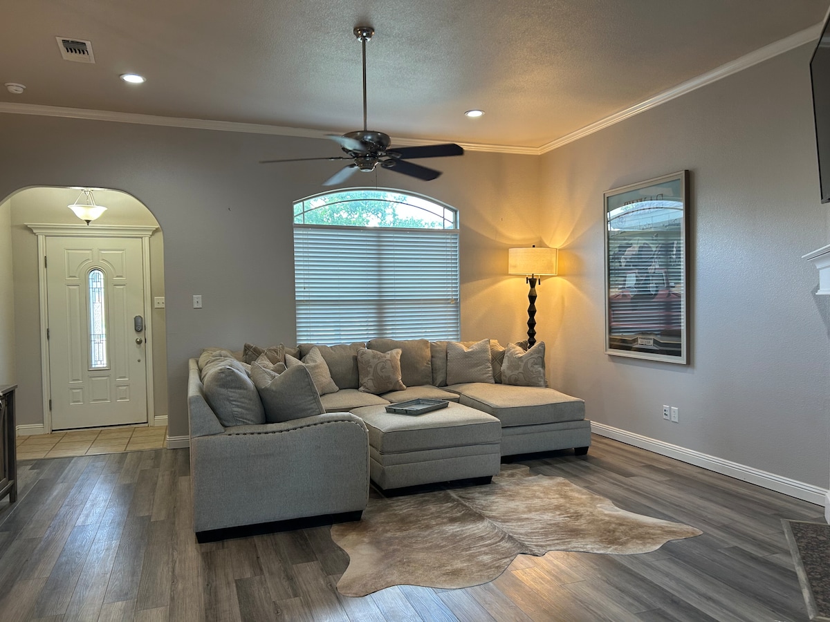 A spacious living area features a comfortable sectional sofa arranged around a small coffee table. Light filters through the large arched windows, and a floor lamp stands beside the sofa. A decorative area rug adds texture to the wooden floor.
