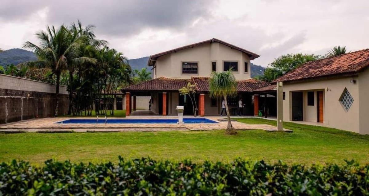 A spacious property is surrounded by lush greenery and features a serene swimming pool. The two-story building, with a traditional roof, is complemented by palm trees and is set against a backdrop of mountains, creating a tranquil outdoor environment.