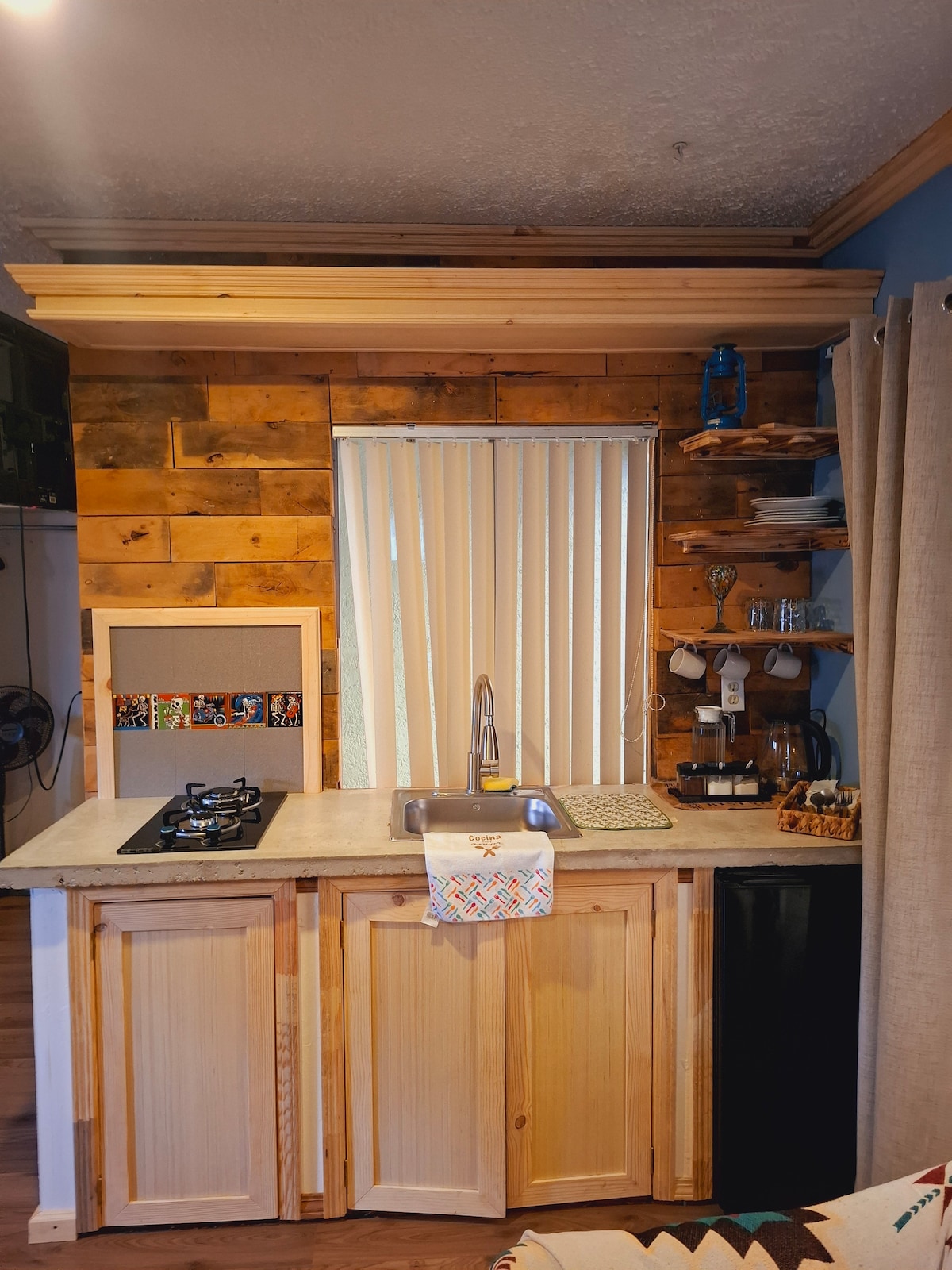 A compact kitchen space is presented, featuring wooden cabinetry and a countertop with a sink. A gas stove is located on one side, while a mini refrigerator stands underneath. Shelves hold kitchen essentials, and blinds cover a window allowing natural light.