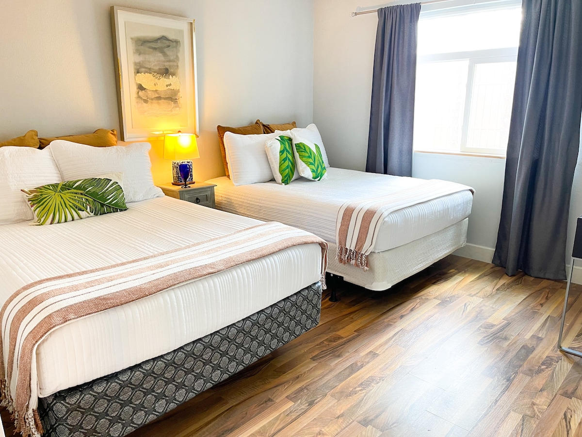 A welcoming bedroom arrangement features two spacious beds adorned with soft white linens and decorative pillows. A side table with a lamp sits between the beds, while natural light streams in through a large window, draped with dark curtains. The room exhibits warm wooden flooring.