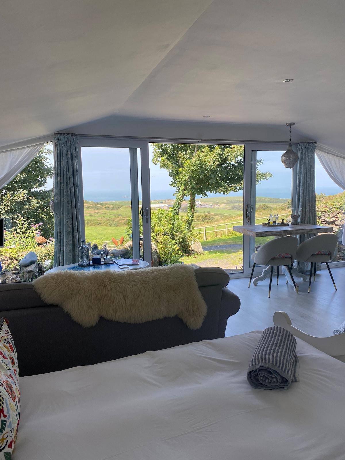 A spacious room features large glass doors leading to an outdoor view of rolling hills and the sea. Sunlight filters through sheer curtains, illuminating a cozy seating area with a fluffy throw. A dining table is placed near the window, enhancing the tranquil ambiance.