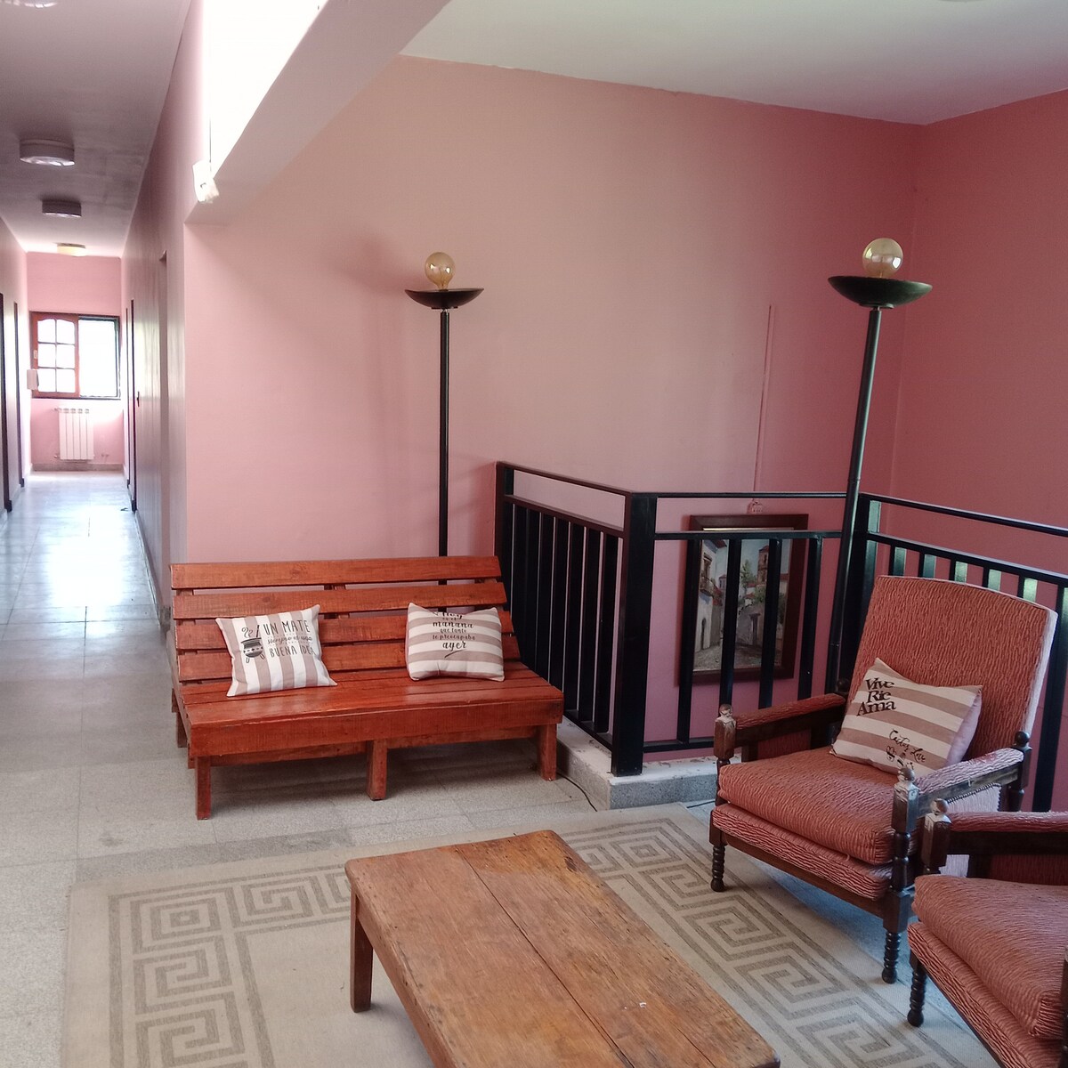 A hallway is lined with smooth, light-colored flooring. Two seating areas feature a wooden bench and an armchair, both adorned with throw pillows. Soft lighting is provided by tall lamps, while pale pink walls create a bright ambiance.