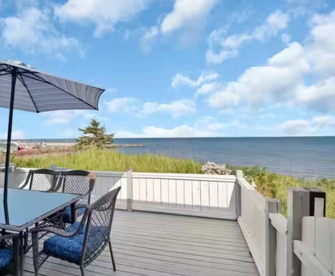 A deck furnished with a table and chairs offers an unobstructed view of the ocean. A large umbrella provides shade, enhancing comfort. Coastal grasses and shrubs frame the edge of the deck, while the shoreline is visible in the distance beneath a partly cloudy sky.
