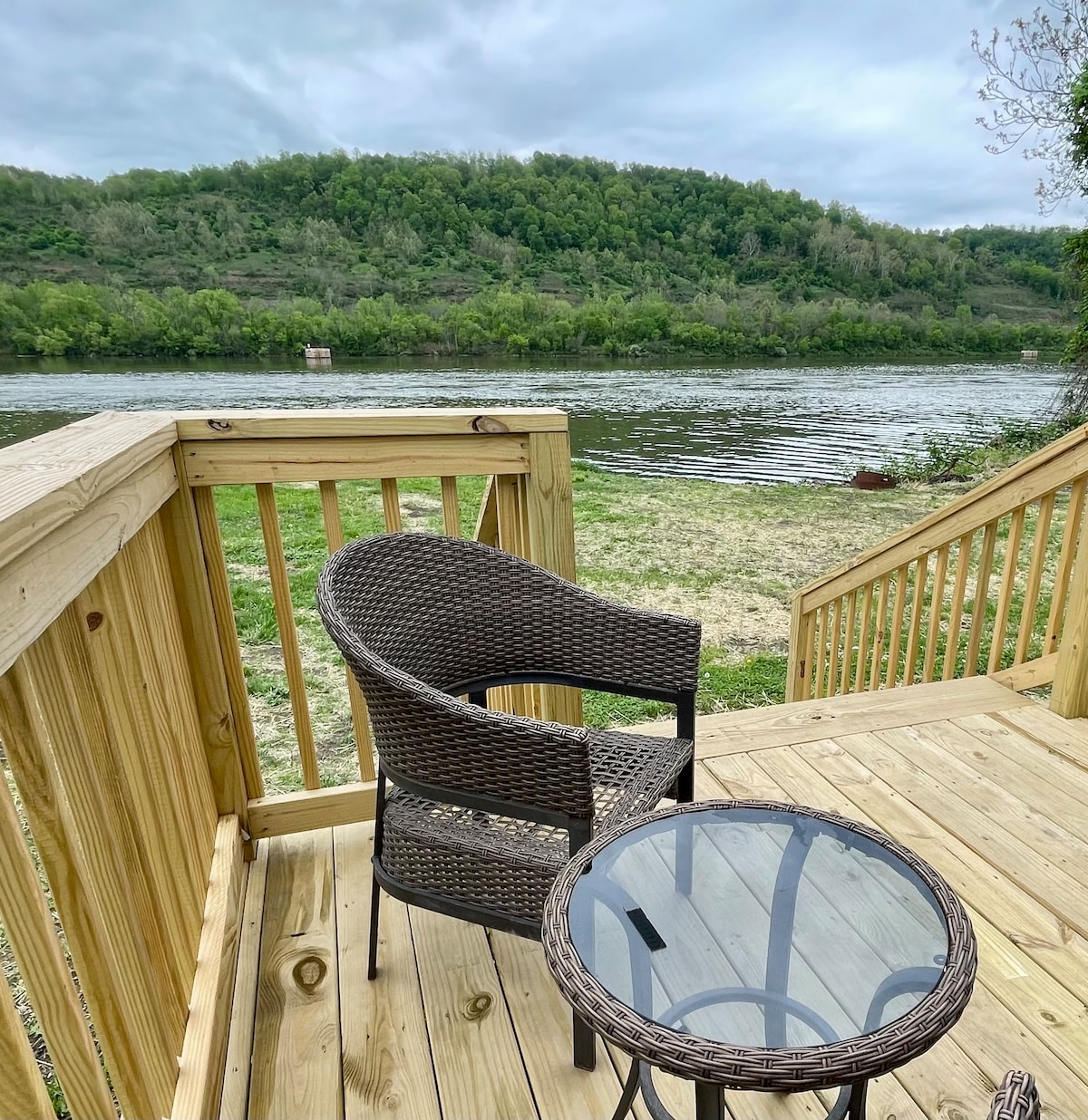 A wooden deck is shown with a small glass table and a woven chair. The Ohio River is visible in the background, surrounded by greenery on the opposite bank, creating a serene riverside atmosphere.