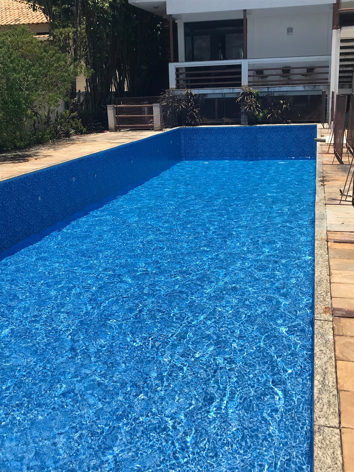 A large swimming pool is surrounded by a paved area, reflecting sunlight on its clear blue surface. The water is inviting, and hints of greenery can be seen in the background, contributing to a peaceful outdoor setting.
