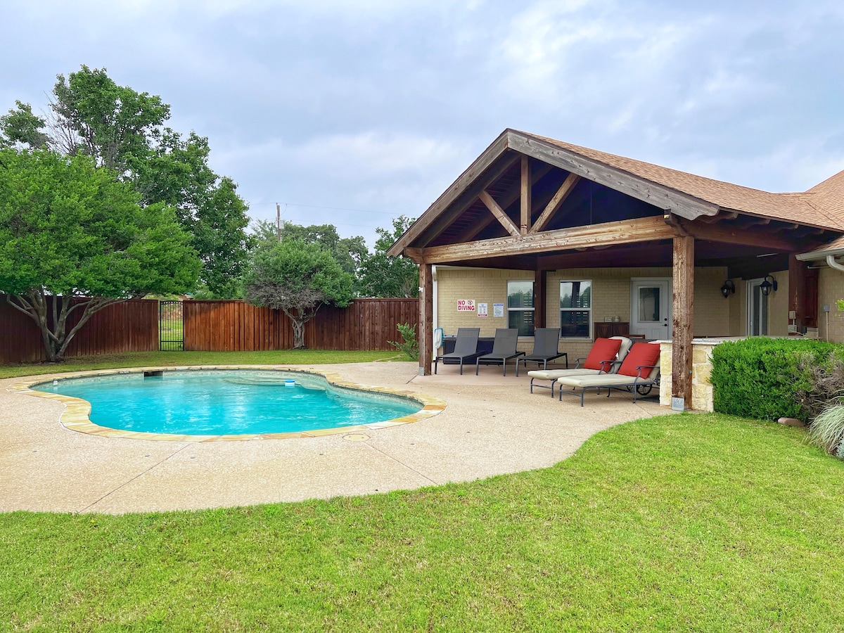A private outdoor space features a shimmering pool surrounded by a concrete patio. Lounge chairs in varying shades line one side, inviting relaxation. Green grass and trees provide a natural setting, while a covered area is visible, offering shade for outdoor gatherings.