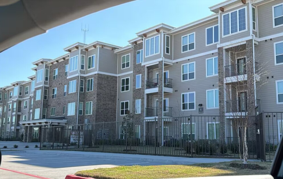 A modern apartment building features three stories with a mix of beige siding and stone accents. Large windows provide natural light, while balconies are present on select units. The gated entry adds a layer of security, framed by landscaped greenery and open space in front.
