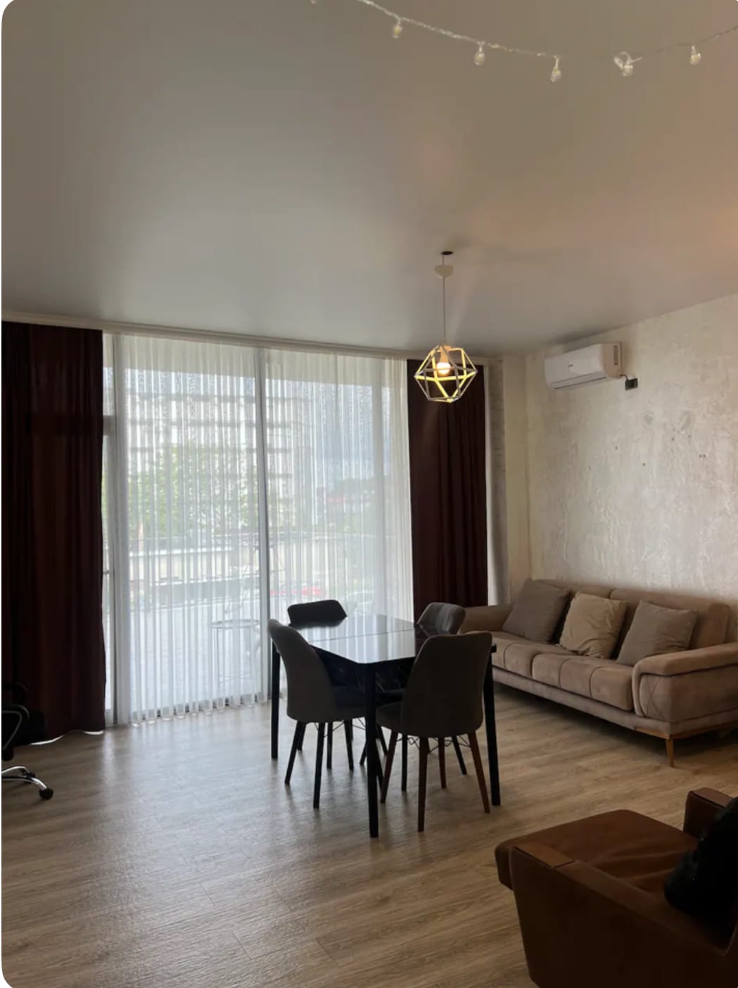 A bright living space features large windows covered by sheer curtains, allowing natural light to fill the room. A comfortable sofa is positioned near the window, accompanied by a dining table with four chairs. A stylish pendant light hangs from the ceiling.