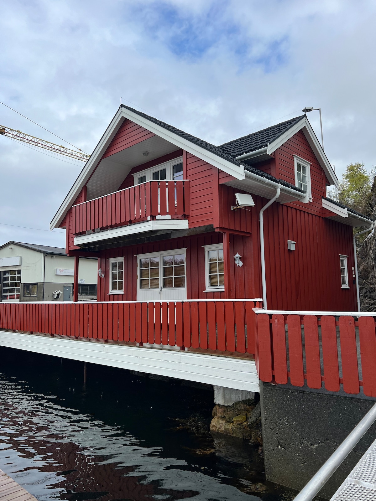 A charming red wooden cabin features a second-story balcony with wooden railings, overlooking the calm waters below. The structure is set against a backdrop of grey skies, while the wide deck extends toward the water, inviting relaxation and scenic views.