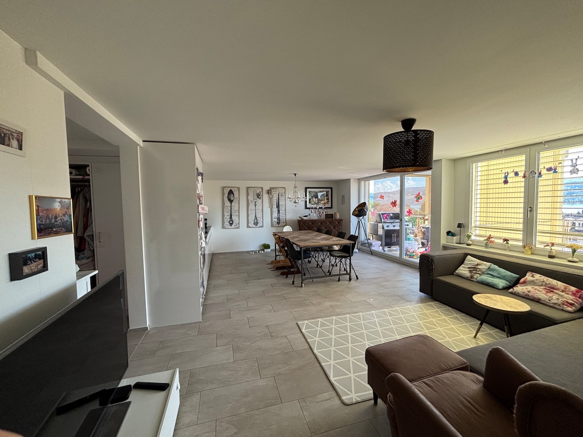 A spacious living area is showcased, featuring a comfortable sofa and a round coffee table. A dining table with several chairs is positioned nearby. Large windows allow natural light to brighten the room, while a balcony view is visible through the glass doors.