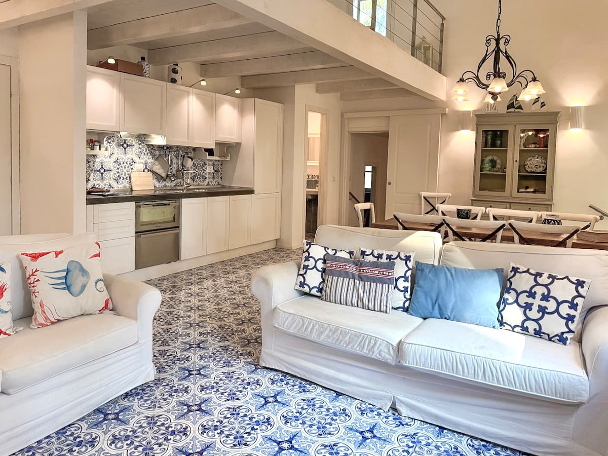 An open living space features a modern kitchen with light cabinetry and intricate blue patterned flooring. Two white sofas are adorned with decorative pillows, creating a comfortable seating area. A dining space is visible in the background, complemented by natural light from large windows.