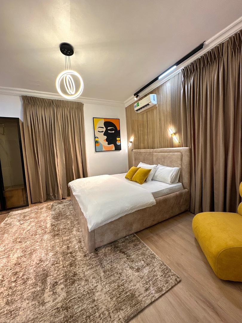 A modern bedroom features a spacious bed adorned with white bedding and decorative yellow pillows. Elegant curtains frame the windows, and a striking wall art piece is displayed above the bed. A cozy chair adds a pop of color, creating a welcoming environment.