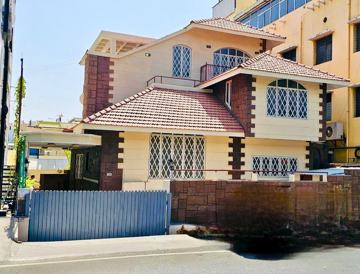 The exterior of a spacious two-story house is showcased, featuring a unique blend of light-colored walls and stone accents. A tiled roof covers the structure, and ornamental ironwork decorates the windows. A gated entrance leads to a private yard, with nearby buildings visible in the background.