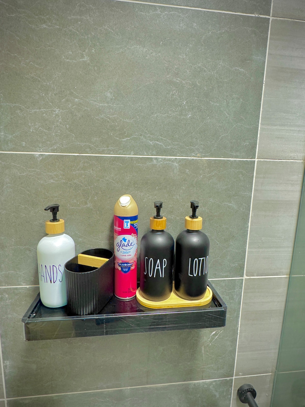 A bathroom shelf displays neatly arranged containers. Two large black bottles labeled 'SOAP' and 'LOTION' sit beside a smaller bottle marked 'HANDS.' A can of air freshener is positioned between the bottles, all set against a backdrop of textured gray tiles.