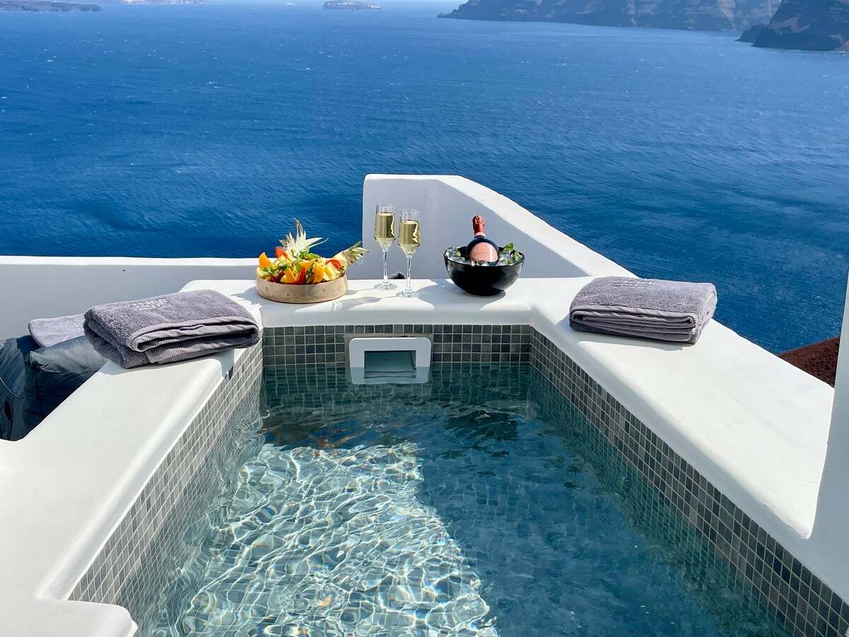 A heated jacuzzi is showcased on a private terrace, overlooking the calm, blue waters of the Aegean Sea. Beside the jacuzzi, a bowl of fresh fruit is presented, alongside two glasses of champagne and a bottle, with soft towels neatly arranged nearby.