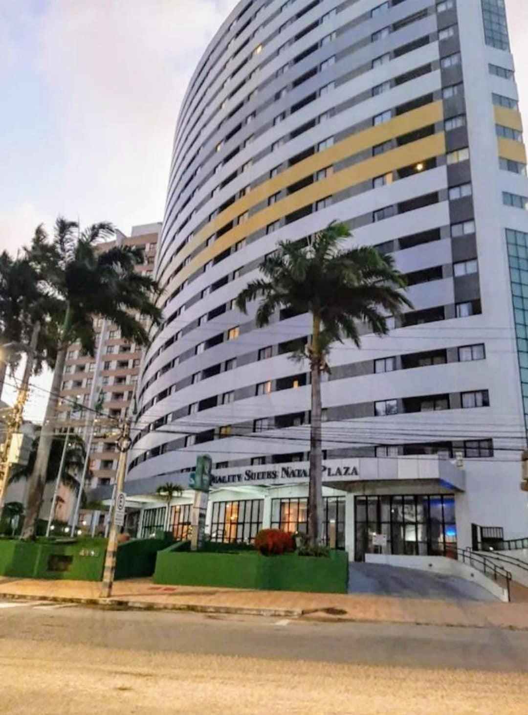 The exterior of the Quality Suites Natal Plaza is presented, showcasing a modern high-rise building with a curved design. Tall palm trees are positioned in front, complemented by landscaping along the base. Large windows reflect the surrounding area, allowing natural light to enhance the façade.