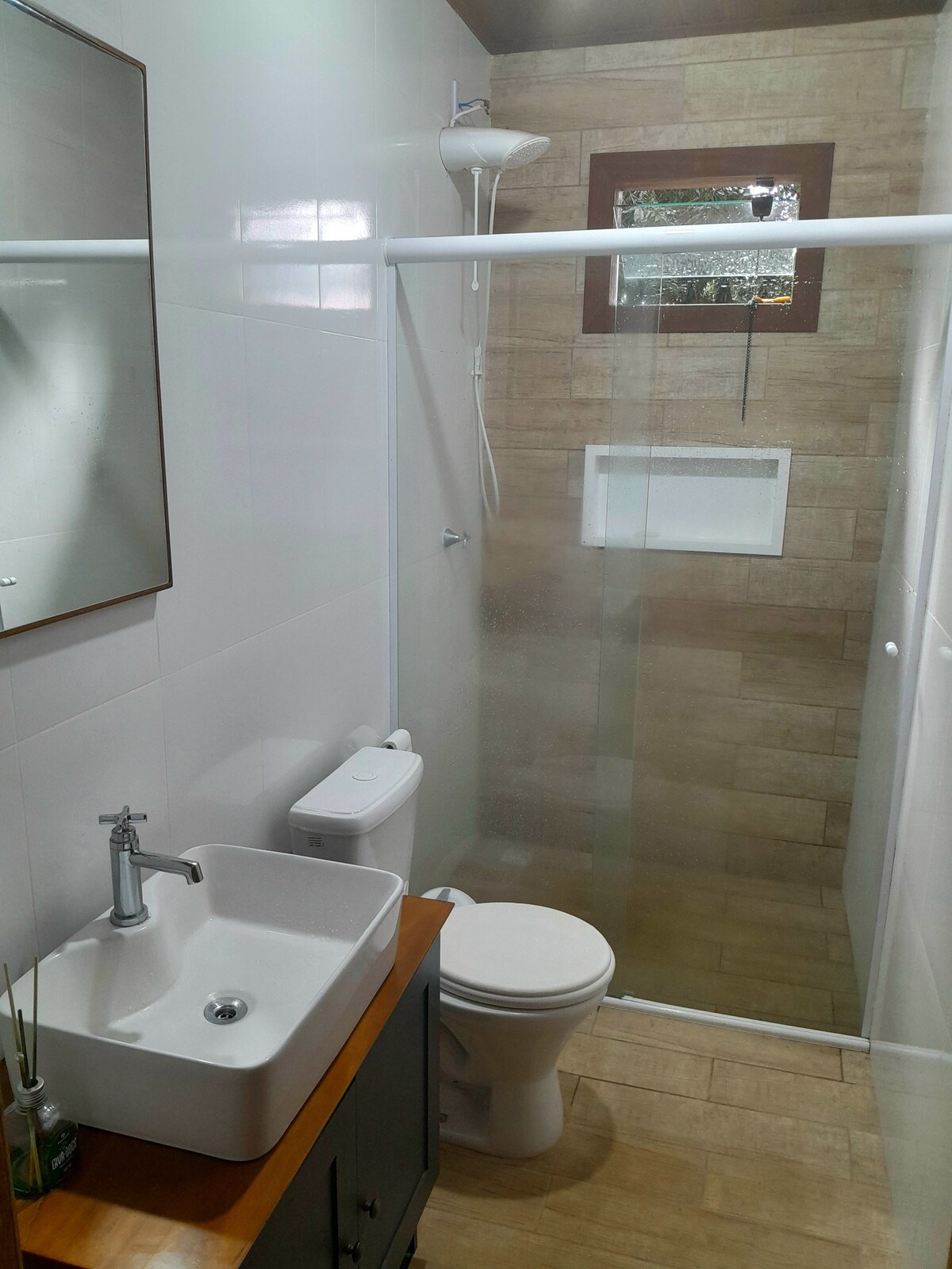 A compact bathroom features a modern sink situated on a wooden countertop. A glass shower enclosure stands alongside, with beige tiled walls creating a neutral backdrop. Natural light is provided through a small window, enhancing the overall functionality of the space.