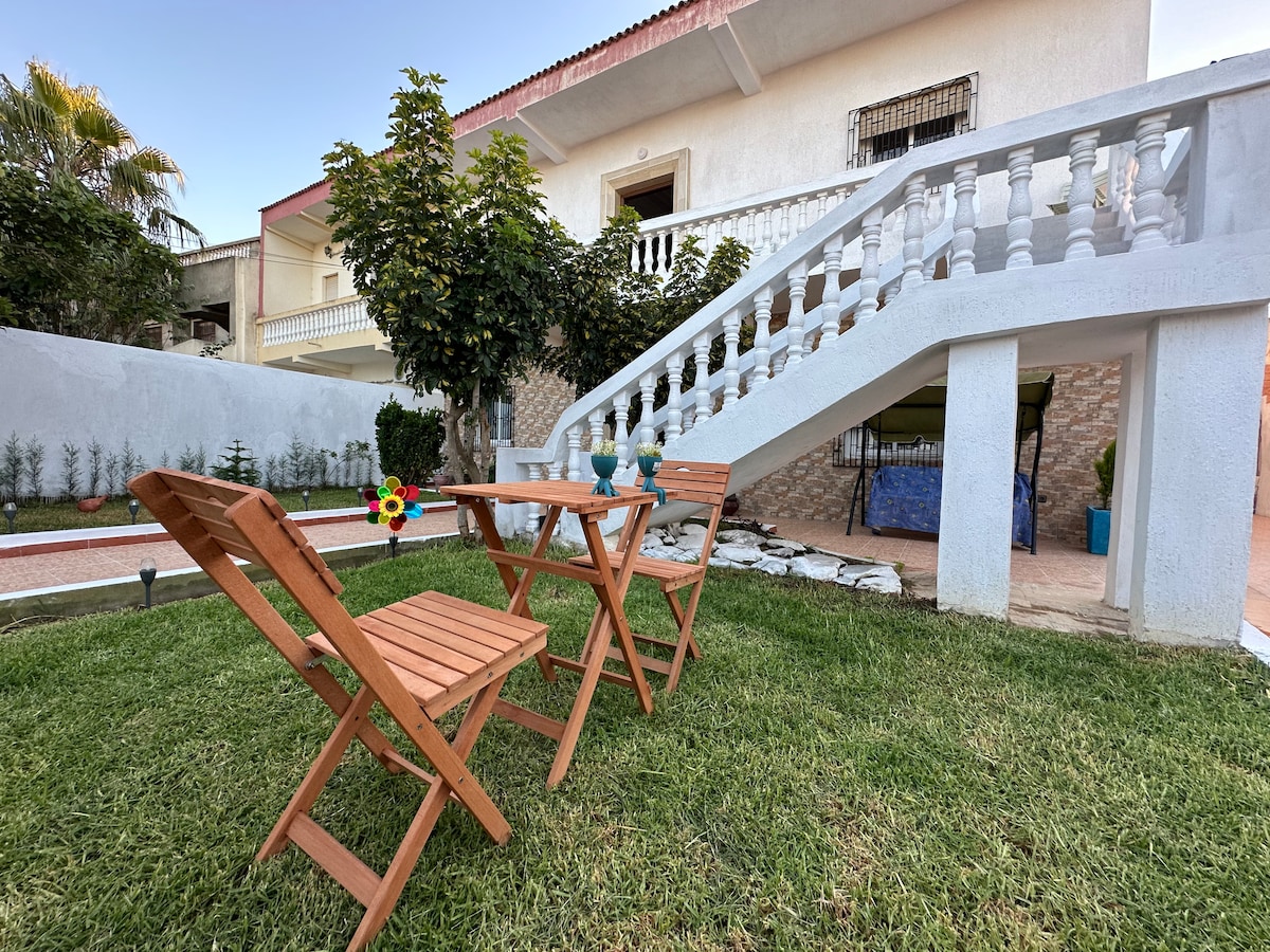 A wooden table and two folding chairs are positioned on a grassy area in the garden. Lush greenery surrounds the space, and a staircase leads to the upper level of the villa. A few potted plants add a touch of color to the inviting outdoor setting.