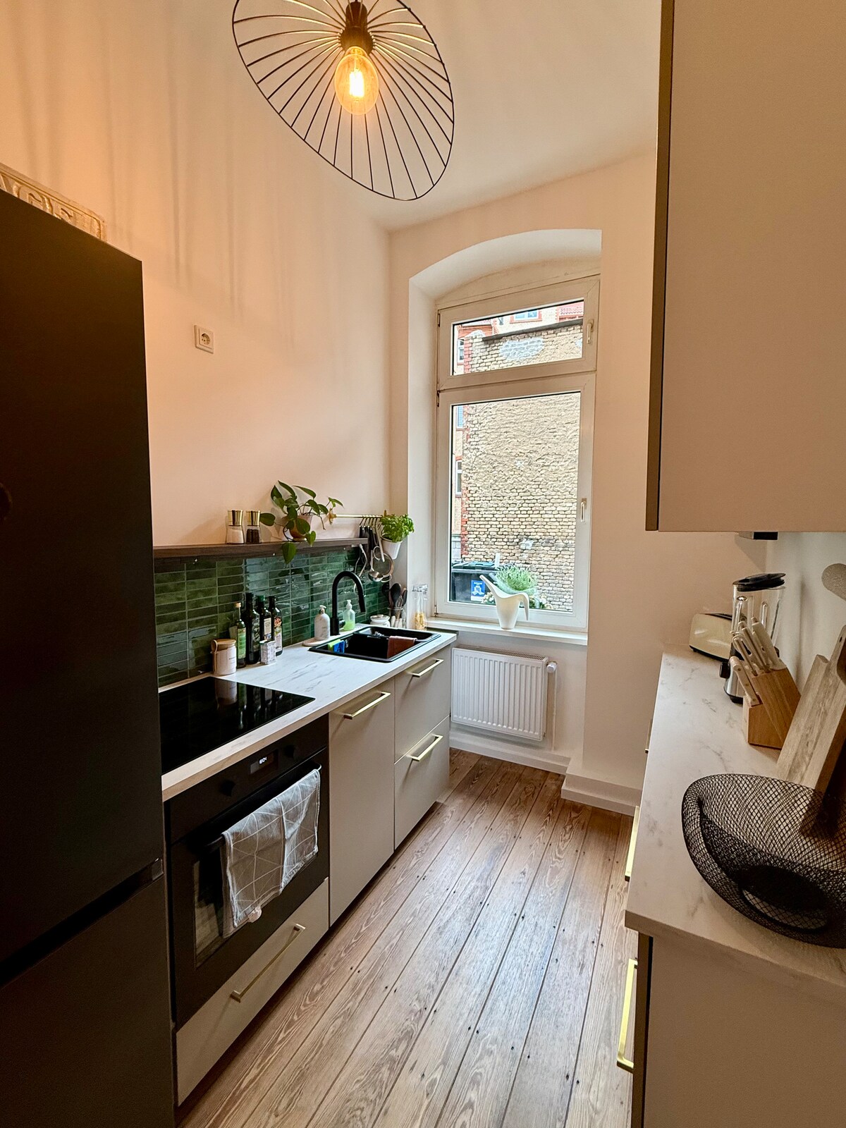 A compact kitchen features modern cabinetry and a sleek countertop. A window allows natural light to brighten the space. Cooking amenities include an oven, stove, and dishwasher. Green tiles add a refreshing touch, while potted plants bring a hint of nature indoors.
