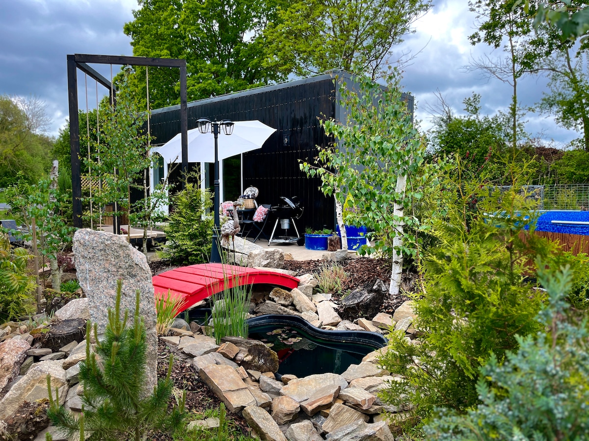 A well-maintained garden area surrounds a stylish black residence, featuring a red wooden bridge over a small pond. Lush greenery, rocks, and colorful planters enhance the peaceful outdoor setting, while outdoor seating and a barbecue grill offer spaces for relaxation and dining.