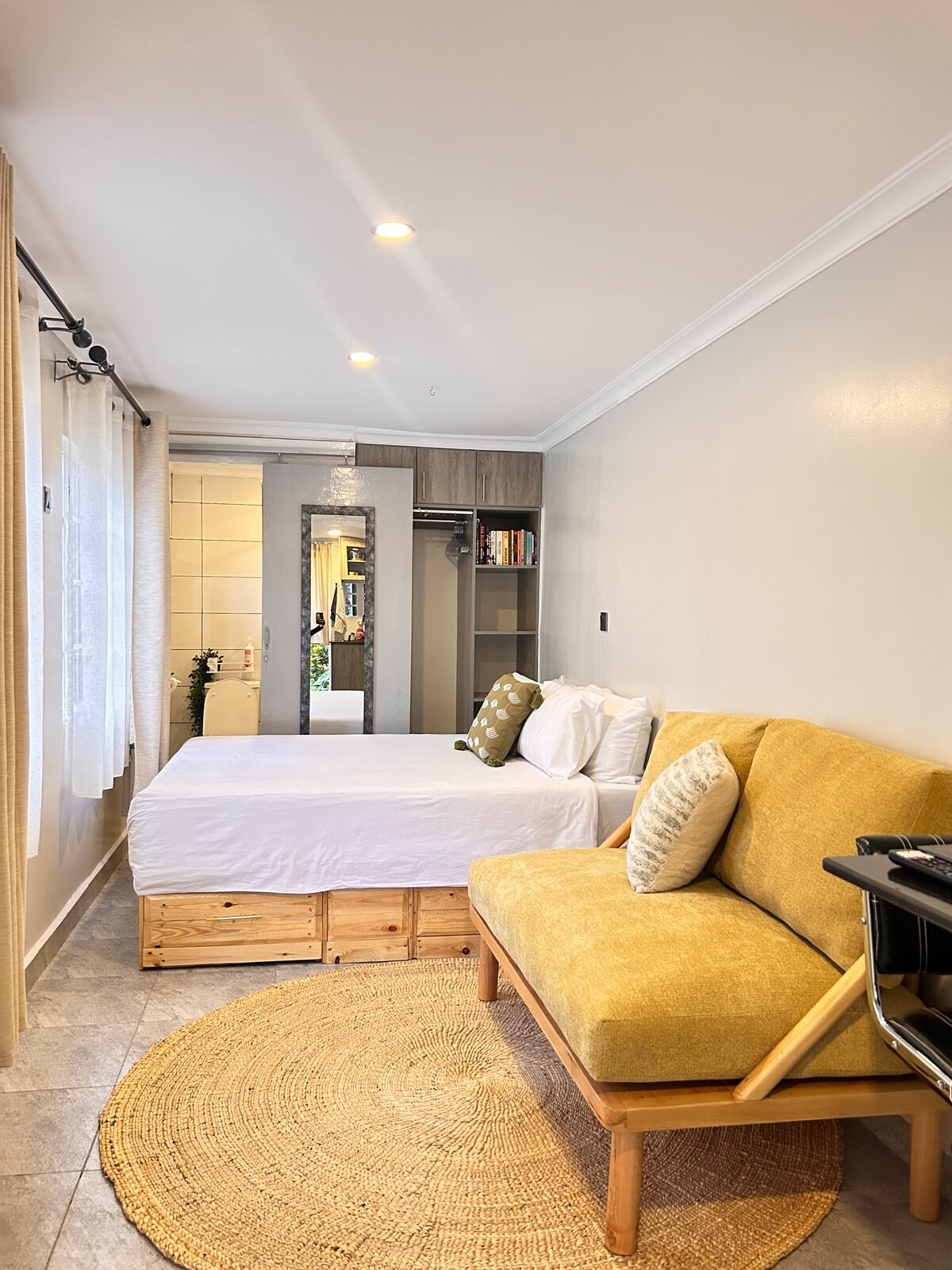 A cozy studio space features a spacious bed with pillows resting against a natural wood base. A bright yellow couch is positioned nearby on a woven rug. Soft natural light enters through sheer curtains, illuminating the minimalistic design elements and layout.