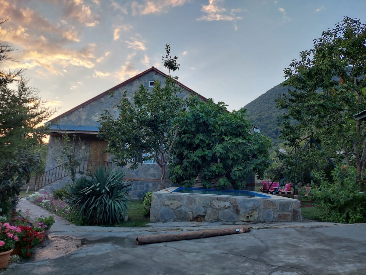 A stone house is surrounded by lush greenery, featuring a small pool in the foreground. Various trees and plants create a natural environment, while a gentle slope leads to a hillside in the background, illuminated by the soft glow of sunset.