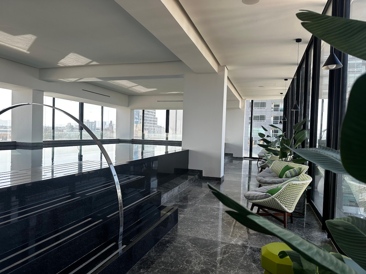 A spacious area features large windows that allow ample natural light. A sleek, modern design is highlighted by polished black stone surfaces and green plants. Comfortable seating arrangements are visible alongside the walls, providing a relaxing atmosphere with a sense of openness.