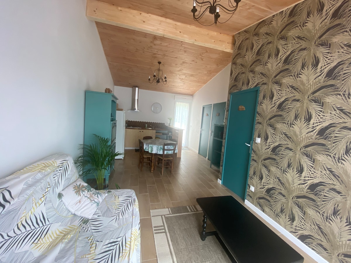 An open living area displays a light-colored sofa adorned with a tropical print. A dining table is set with chairs, while a kitchen area is visible in the background. Natural light fills the space through large windows, complemented by a textured wallpaper featuring a palm leaf design.