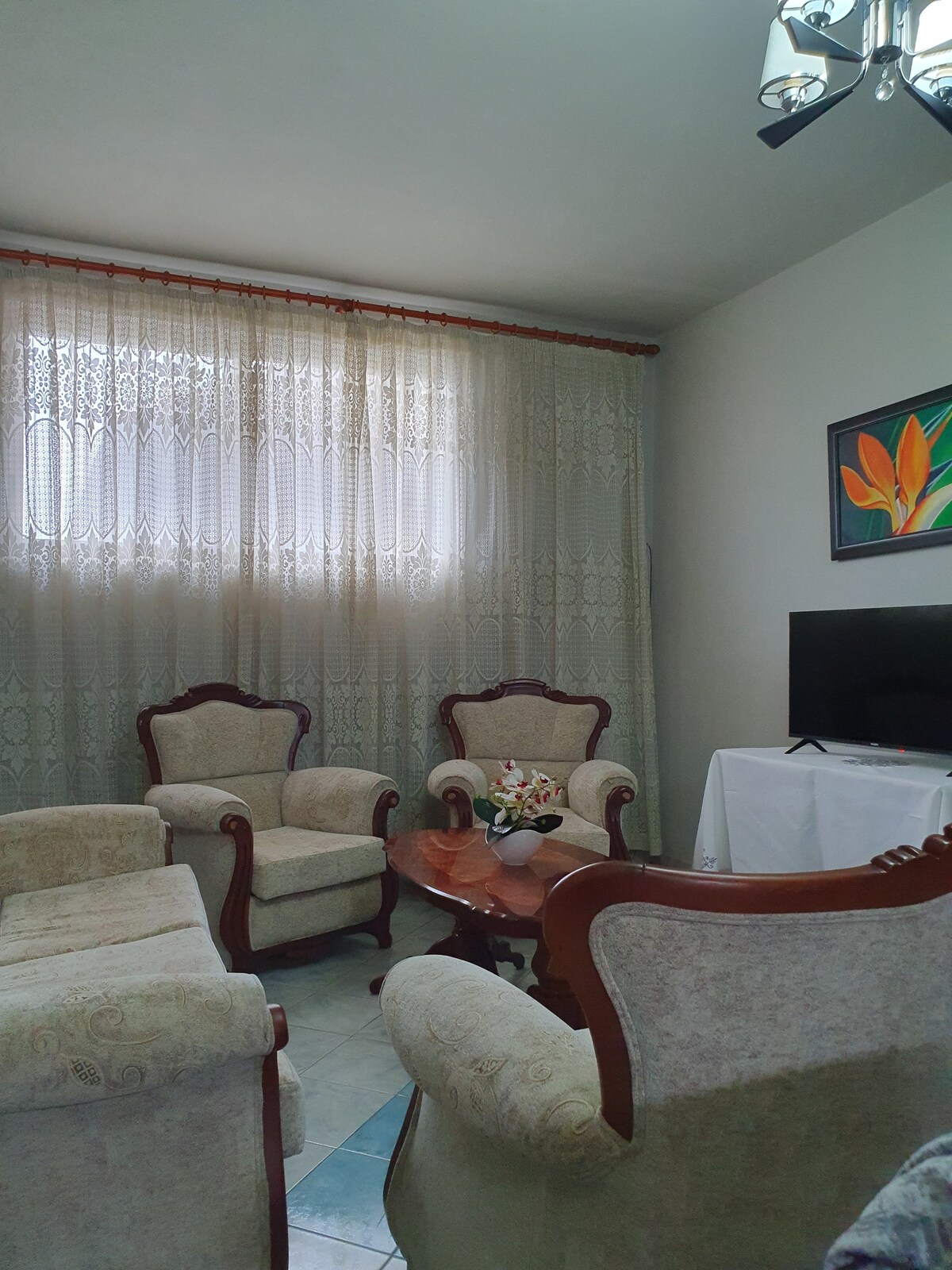 A comfortable living area features two armchairs and a small wooden table, arranged together under a ceiling fan. Natural light enters through lace curtains, illuminating a vibrant piece of artwork on the wall. A television is positioned on a stand in the corner.