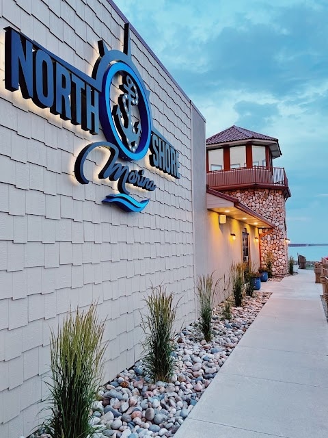 The exterior of North Shore Marina is visible, featuring the prominent signage. A pathway lined with smooth stones and ornamental grasses leads towards the entrance. The structure showcases a blend of traditional and modern architectural elements, with a rooftop tower and large windows overlooking the lake.