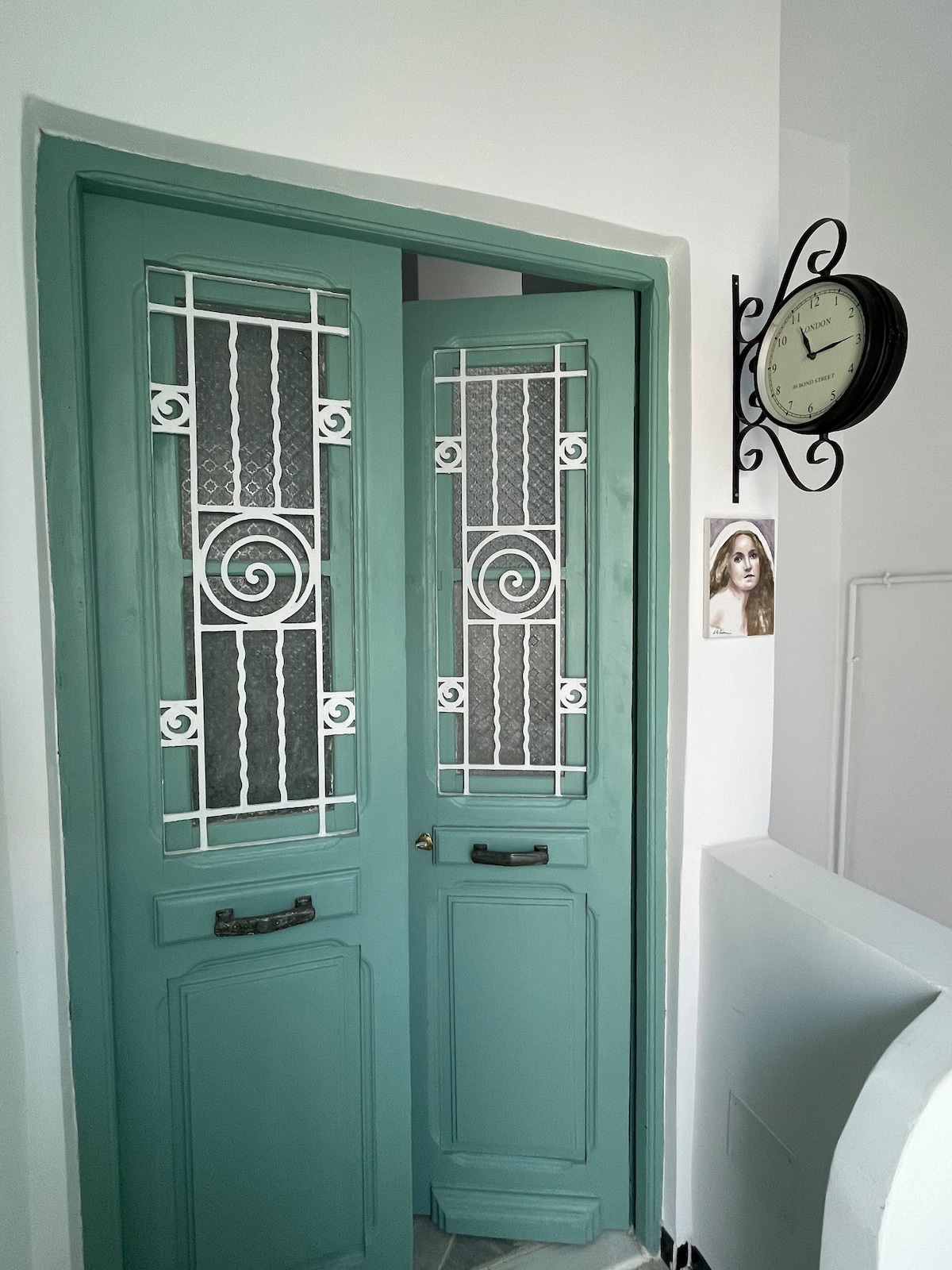 The image shows a set of elegant double doors in a soft turquoise color, featuring decorative ironwork. A classic wall clock is mounted nearby, indicating the passage of time. The surroundings are bright and minimalist, creating a welcoming entryway.