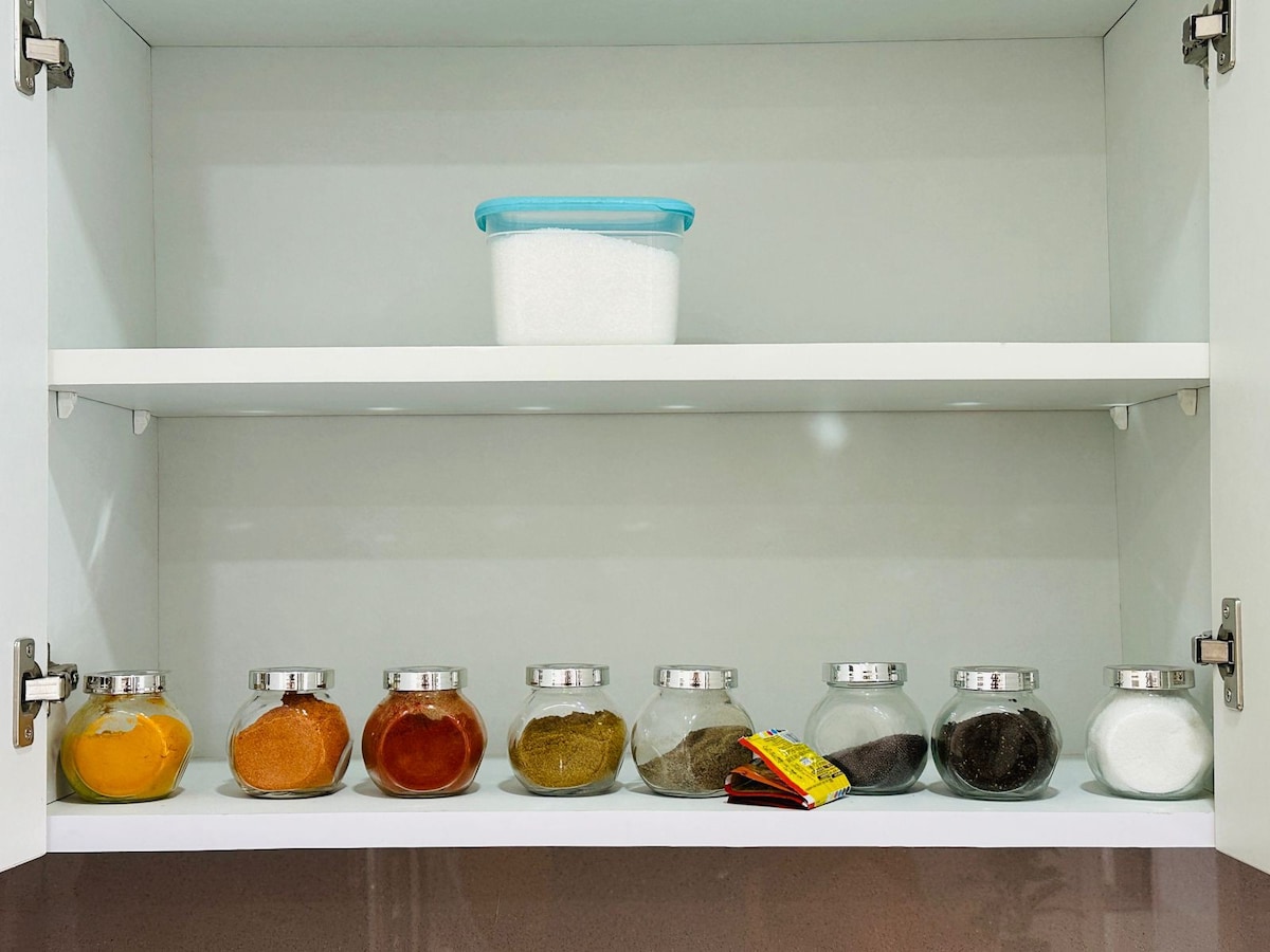 An organized kitchen cabinet is shown, featuring two shelves. The lower shelf displays eight glass jars filled with various spices, each with a silver lid. A container of sugar is placed on the top shelf, enhancing the functionality of the space.