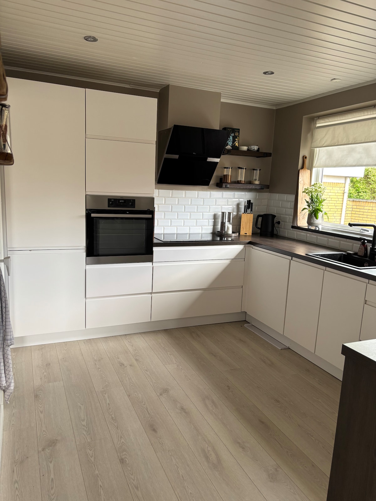 A modern kitchen is featured, showcasing white cabinets and a sleek black oven. A light tile backsplash complements the space, while natural light filters in through a window with a light shade. Wood-like flooring adds warmth to the overall design.