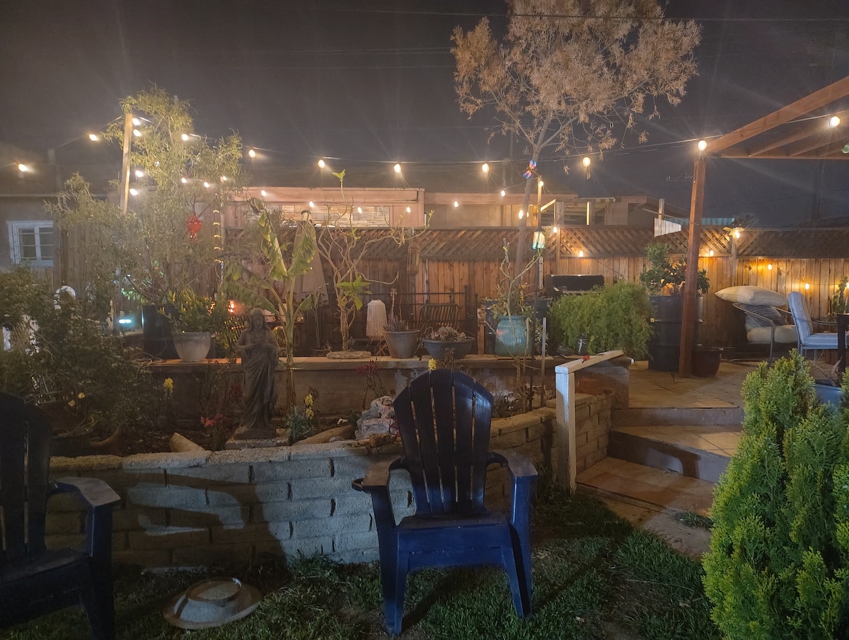 The garden space is illuminated by string lights, creating a soft glow among the various plants and trees. Two blue chairs are positioned nearby, inviting relaxation, while a gravel pathway leads to a wooden deck within the serene outdoor area.
