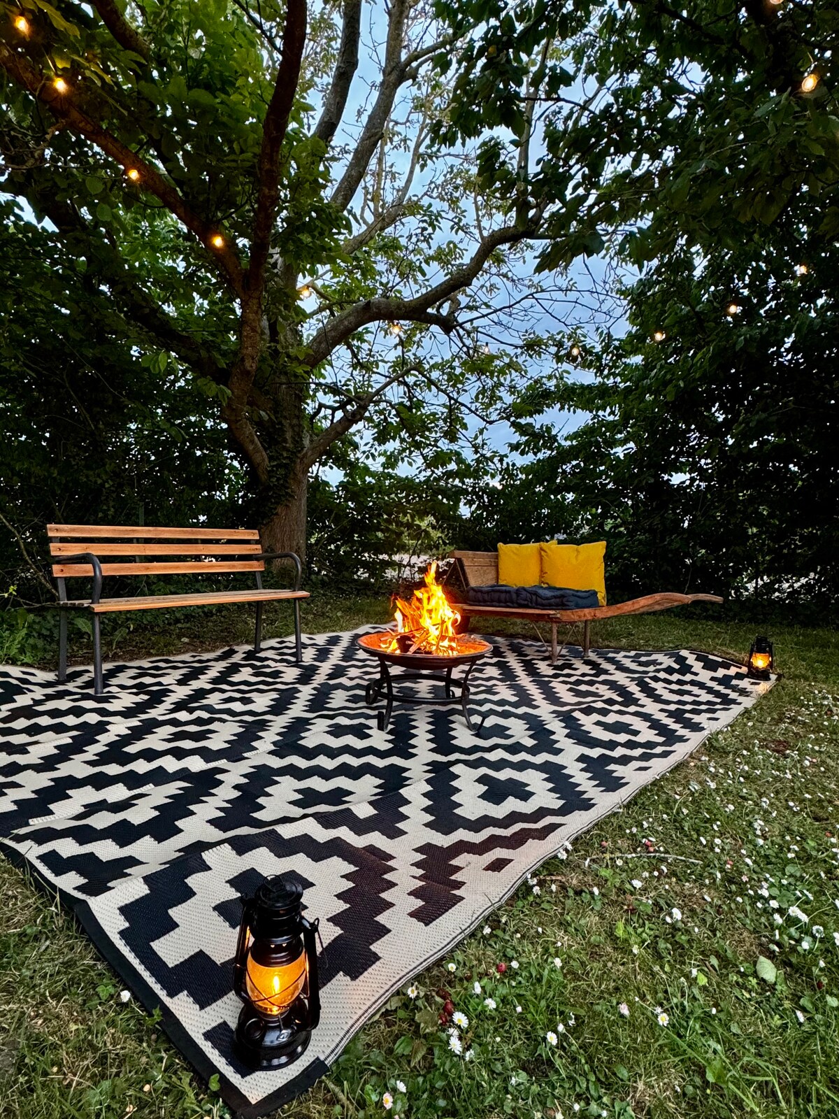 An outdoor seating area is set beneath a tree, featuring a patterned rug, a wooden bench, and a sun lounger with yellow cushions. A fire pit sits in the center, surrounded by lanterns and greenery, creating a cozy atmosphere for relaxation.