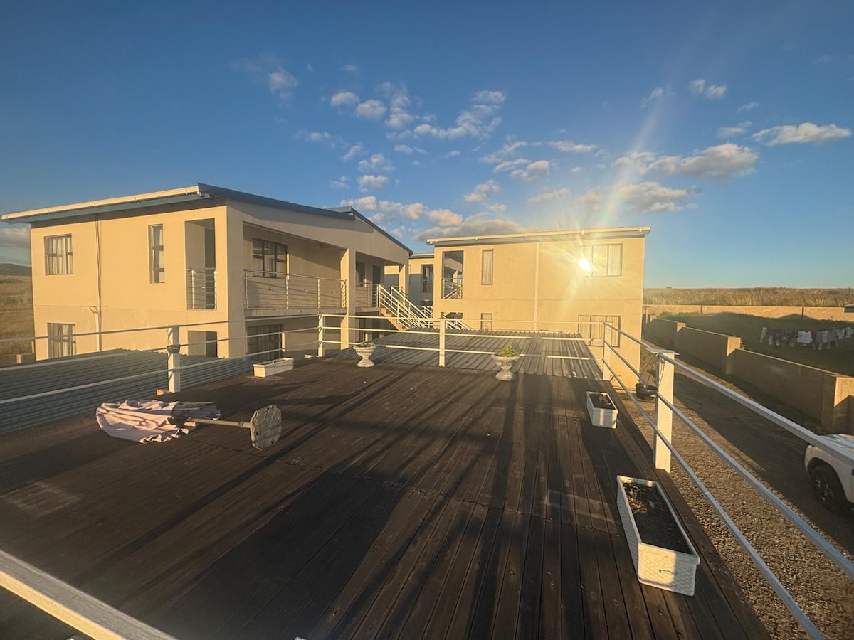 A spacious rooftop deck is featured, showcasing a mixture of potted plants and patio furniture. Natural light reflects off the nearby buildings, while a clear blue sky and soft clouds create a serene backdrop.