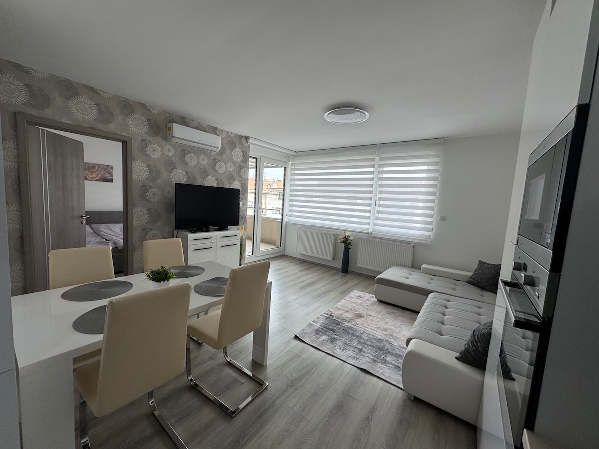 A bright living area features a spacious white sectional sofa and a modern dining table with four upholstered chairs. Large windows are adorned with light-filtering blinds, allowing ample natural light to fill the room. Subtle wall decor complements the contemporary design.
