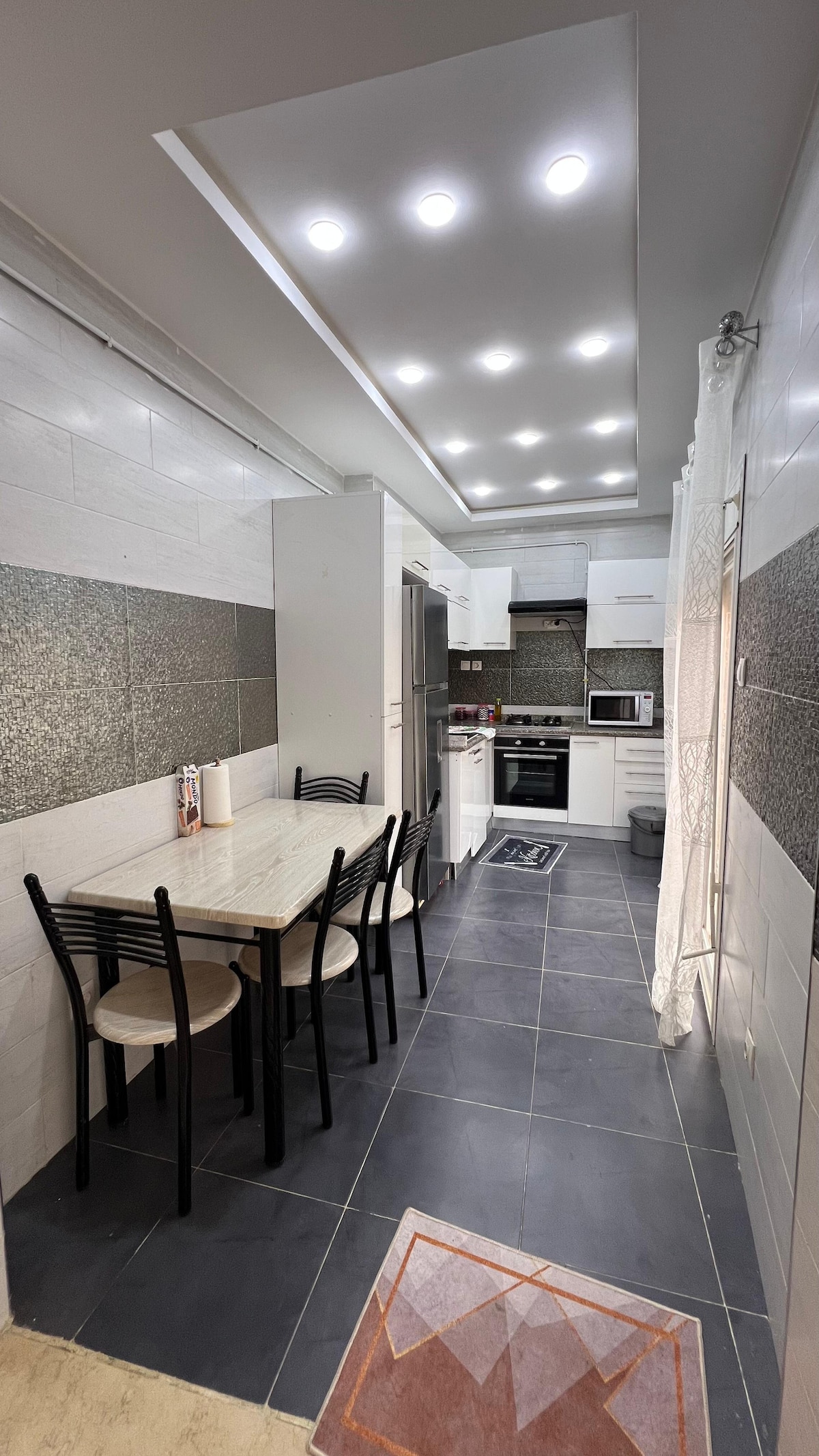 A well-equipped kitchen features a rectangular dining table with six black chairs, offering a functional space for meals. Modern appliances, including a refrigerator and stove, are integrated into sleek cabinetry. Bright overhead lights illuminate the area, enhancing the clean lines and contemporary design.