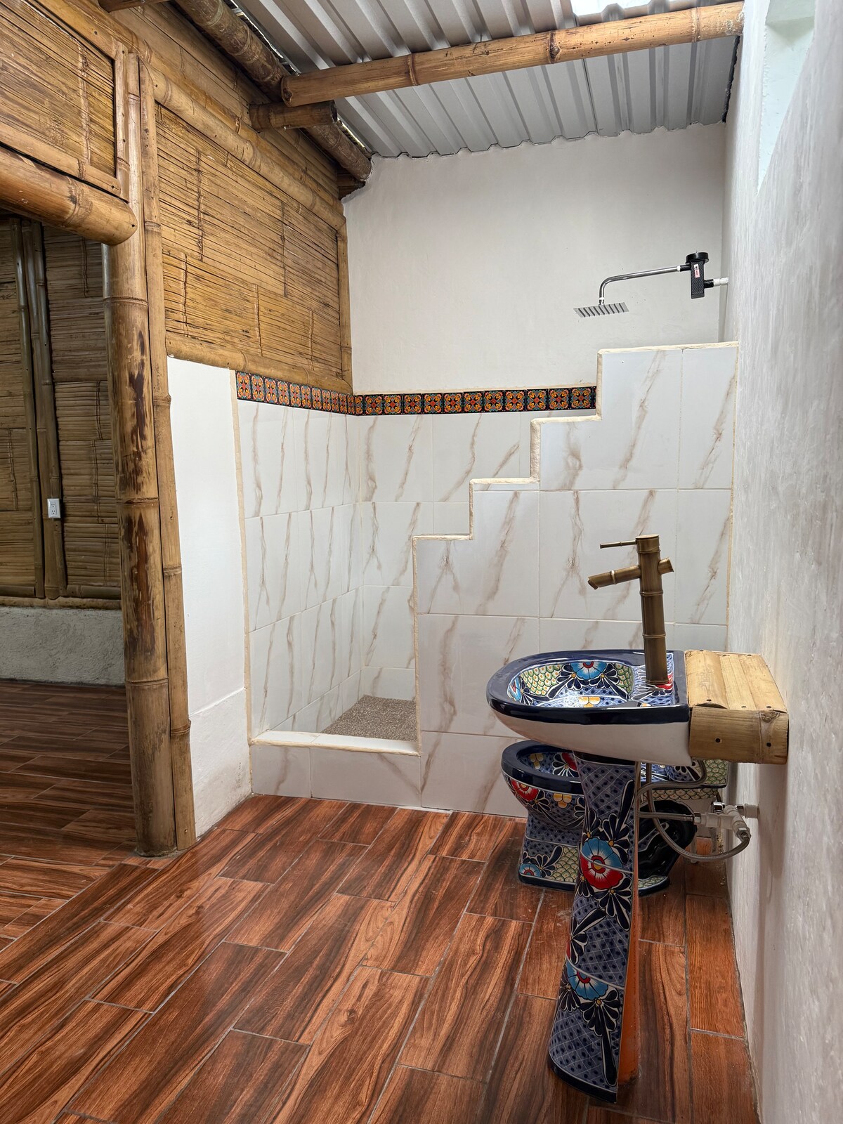 A modern bathroom features a walk-in shower with a simple showerhead, surrounded by elegant tiled walls. A uniquely designed sink with artistic patterns rests on a wooden platform. Natural light filters through a nearby window, highlighting the bamboo accents and warm wooden flooring.
