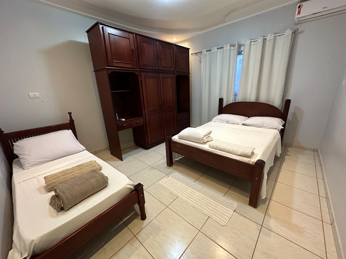 A well-organized sleeping area is equipped with two wooden beds, one larger and one smaller. Fresh linens, towels, and blankets are neatly arranged on each bed. A large dark wood wardrobe with shelving is positioned against the wall, while a window dressed with light curtains allows natural light to filter in.