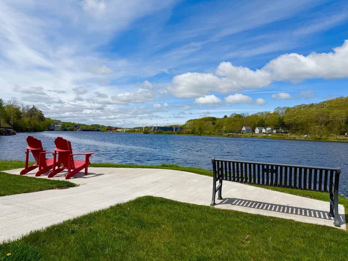 A scenic waterfront view features two red Adirondack chairs positioned beside a pathway, overlooking a calm river. A black bench is also visible, situated on the grassy area. Lush green trees line the opposite bank, under a blue sky with scattered clouds.