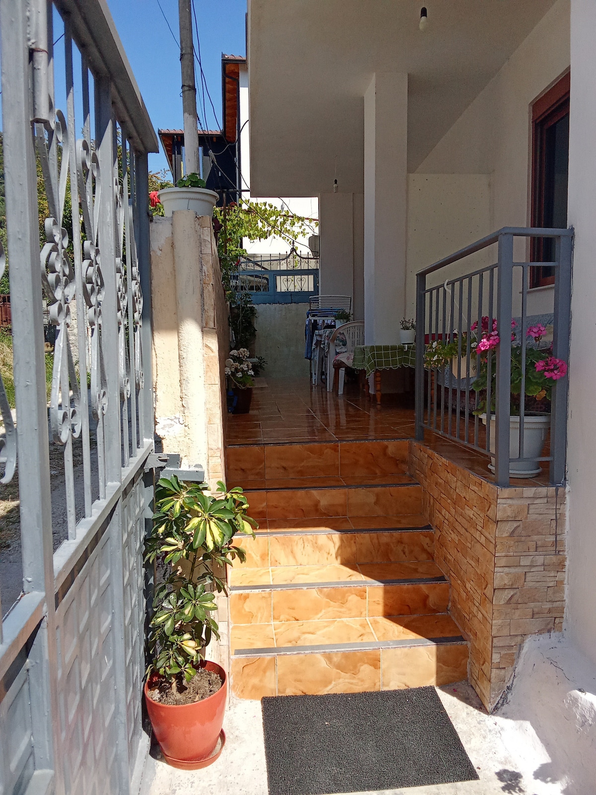 A welcoming entrance features a set of stairs leading up to an open porch area. Potted plants adorn the stairway, while a seated area with a table is visible in the background. Natural light illuminates the space, enhancing its inviting appearance.