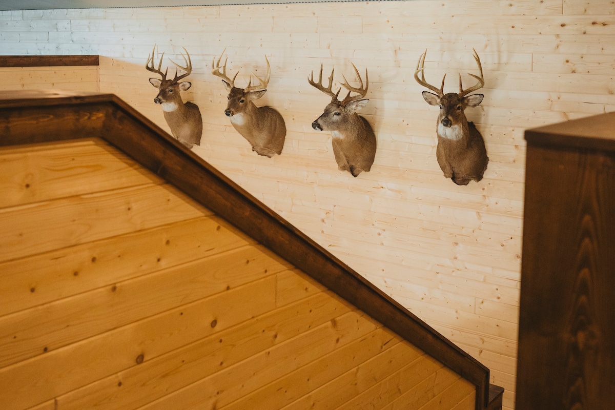 The staircase features wooden steps leading upward, with a natural wood paneling backdrop. Four lifelike deer mounts are displayed along the wall, showcasing their antlers and detailed features, contributing to the rustic charm of the entryway.